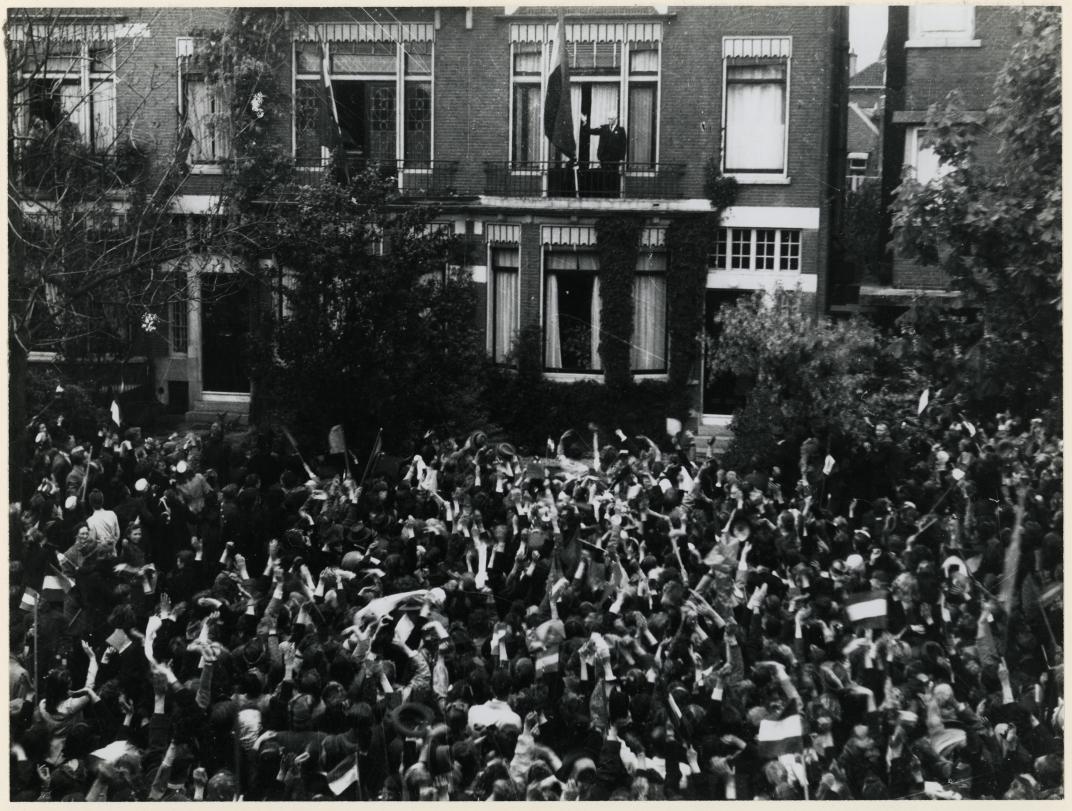 Burgemeester Oud hangt op het balkon van zijn woning aan de Hoflaan de Nederlandse vlag uit. Een grote groep Rotterdammers juicht hem toe en viert de bevrijding van Nederland en het einde van de Tweede Wereldoorlog. Fotograaf Bob van Rhijn, 6 mei 1945, Collectie 4282, 1977-3577.