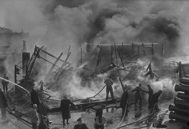 Brandweerlieden blussen de smeulende restanten van een ingestorte gemeenteloods aan de Linker Veerdam. Op de achtergrond grote rookwolken en stapels met buizen; links op de voorgrond twee hoofdmannen van de brandweer, 26-4-1956. Collectie 4147, documentnu