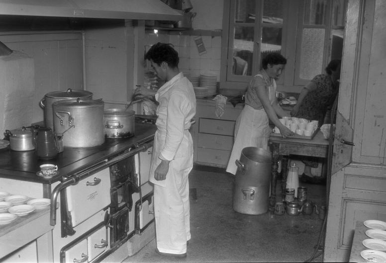 In een keuken, waarschijnlijk op het terrein van de Koninklijke Rotterdamsche Lloyd aan de Lloydstraat, bereiden gerepatrieerde mensen uit het voormalige Nederlands-Indië een maaltijd. Links een fornuis met ovens en grote pannen.